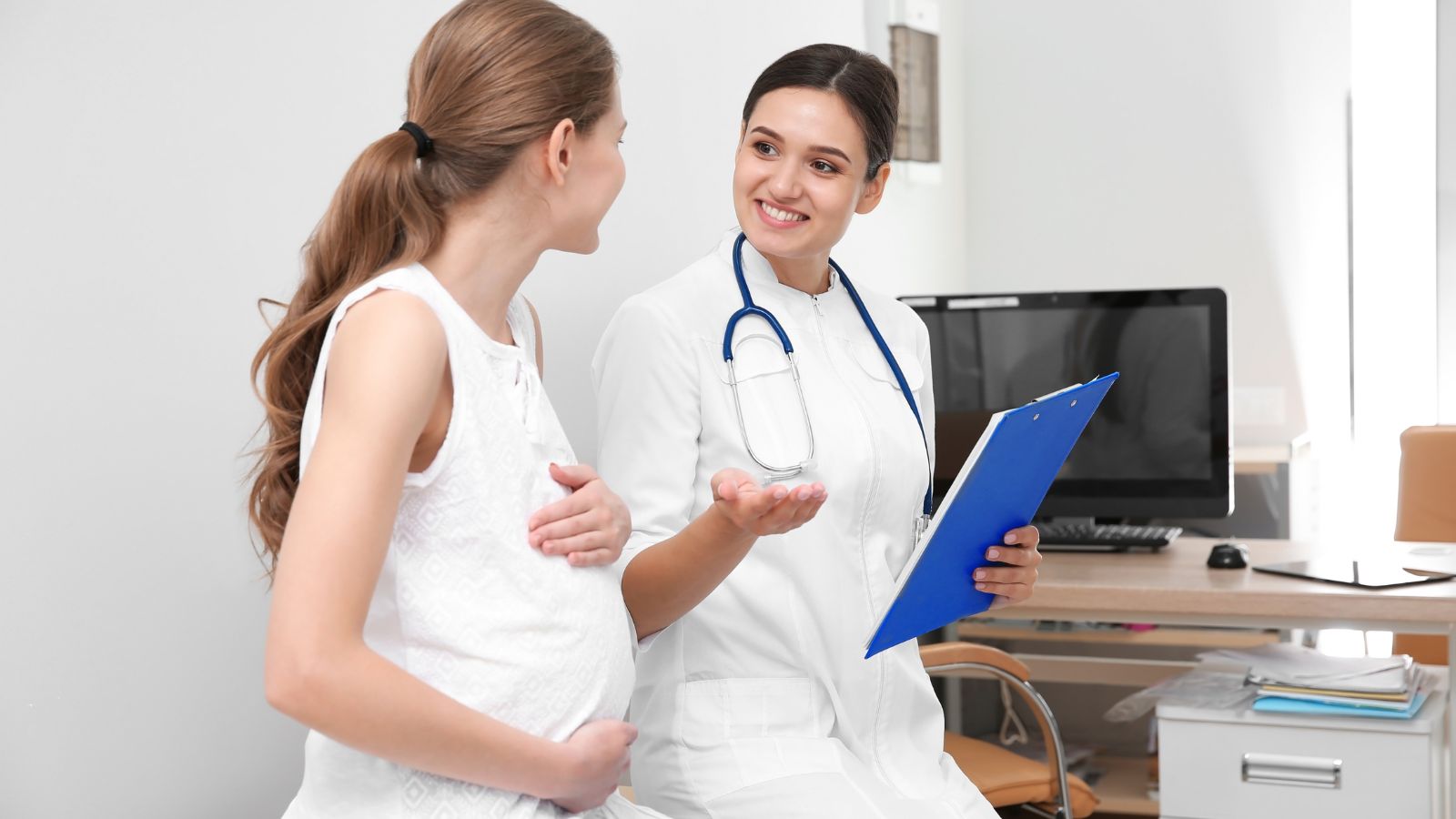Pred začatím cvičenia vždy vyhľadajte gynekologickú konzultáciu. Váš lekár/lekárka či pôrodná asistentka, pozná váš zdravotný stav najlepšie. Zdroj foto: Canva Tehotná žena na konzultácii so svojou lekárkou v ambulancii. Ženy vedú príjemný rozhovor v uvoľnenej atmosfére s úsmevom na perách.